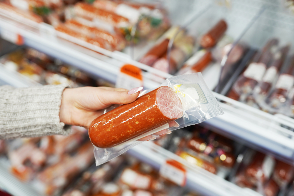 A woman's manicured hand holding a vacuum packages half sausage in a grocery store. 